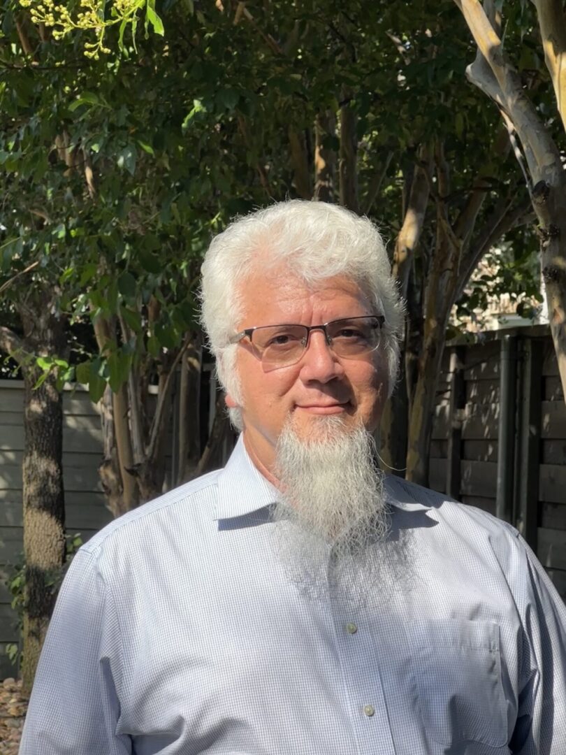 A man with white hair and glasses is standing in front of trees.