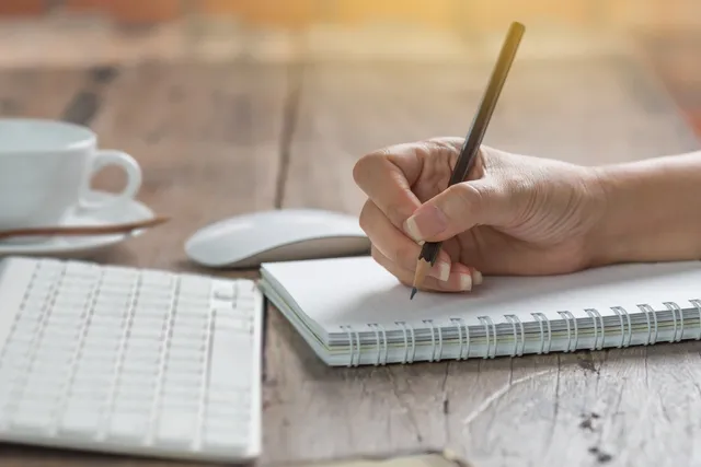 A person writing in a notebook on top of a desk.